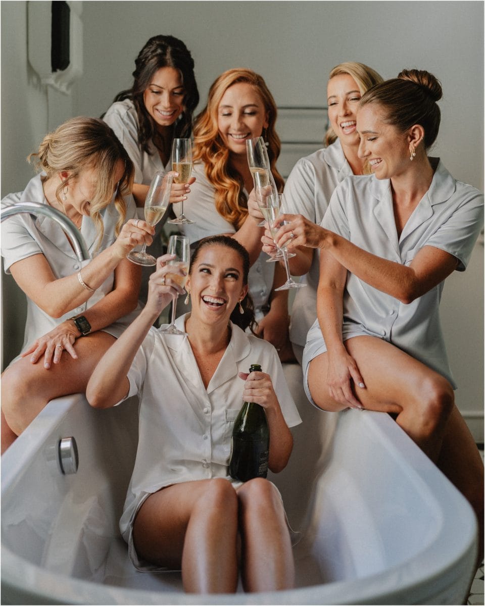 A bride sits in a bathtub drinking champagne with her bridesmaids around her at her Mona Farm wedding